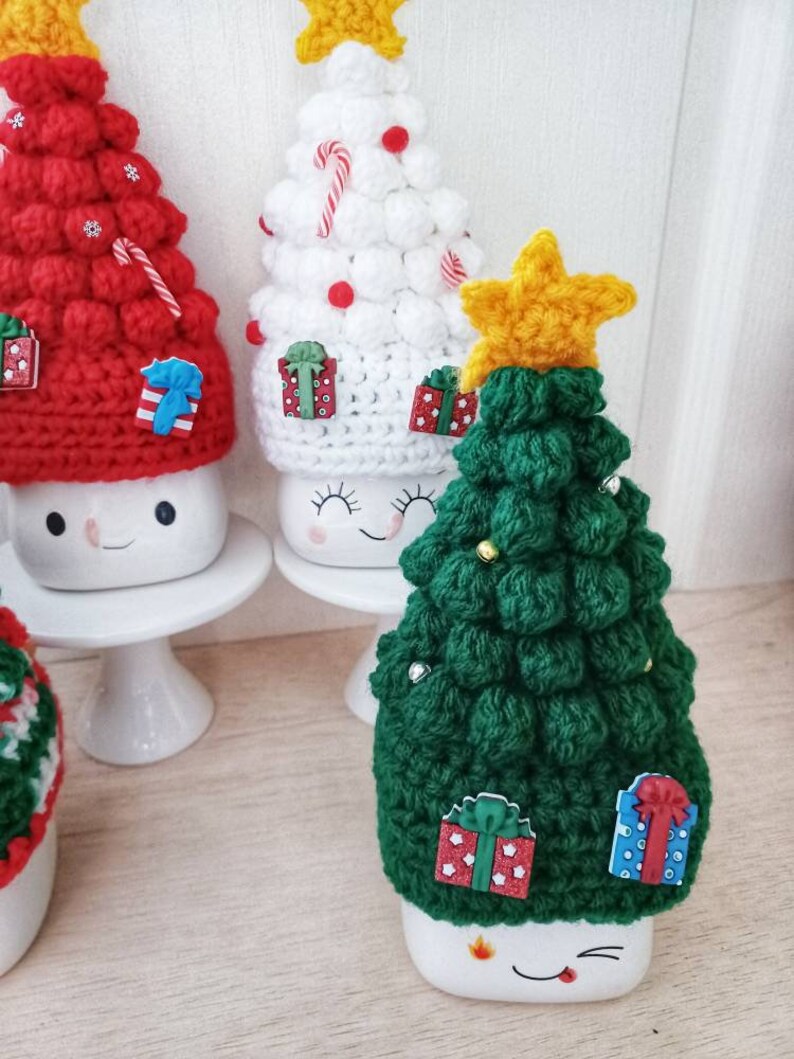 May include: Three crocheted Christmas tree hats in red, white, and green. Each hat has a star on top and is decorated with candy canes, presents, and beads. The hats are sitting on top of white mugs with cute faces.