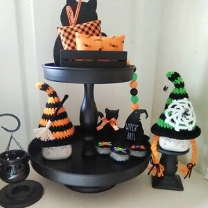 May include: A black tiered tray decorated for Halloween with black cats, witch hats, and a cauldron. The top tier has a black cat silhouette with a plaid pillow and a pillow with a spiderweb pattern. The middle tier has a black cat with a bow, a witch hat with the text "Witch Brew", and two black cats with green and purple bows. The bottom tier has a witch gnome with a black and orange striped hat and a witch gnome with a green and white crocheted hat.