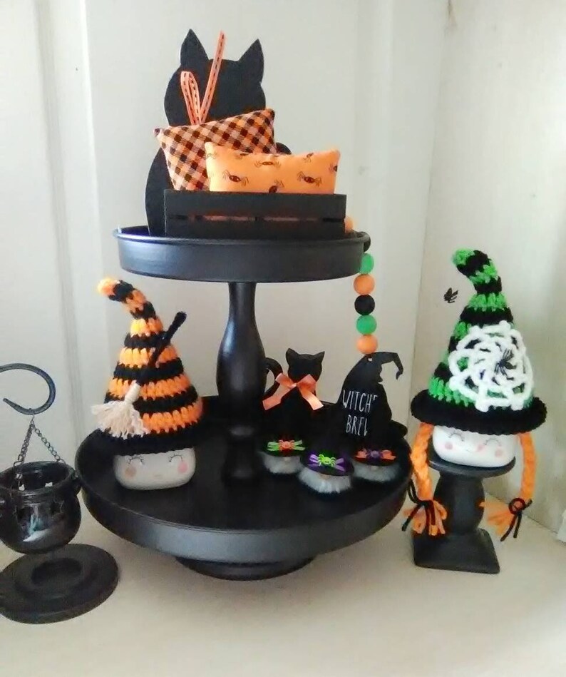 May include: A black tiered tray decorated for Halloween with black cats, witch hats, and a cauldron. The top tier has a black cat silhouette with a plaid pillow and a pillow with a spiderweb pattern. The middle tier has a black cat with a bow, a witch hat with the text "Witch Brew", and two black cats with green and purple bows. The bottom tier has a witch gnome with a black and orange striped hat and a witch gnome with a green and white crocheted hat.