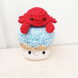 May include: A crocheted blue and brown mushroom with a red crocheted crab on top. The mushroom has a white base with a smiling face.