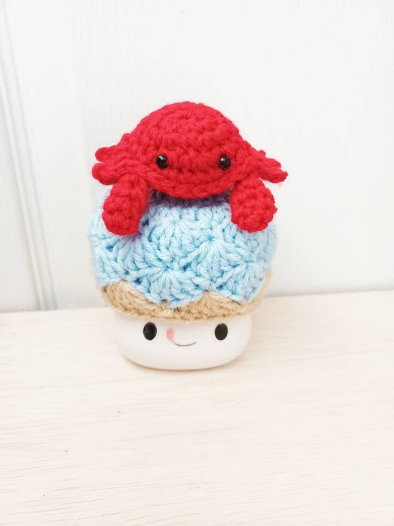 May include: A crocheted blue and brown mushroom with a red crocheted crab on top. The mushroom has a white base with a smiling face.