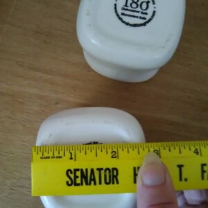 May include: Two white ceramic square bowls with rounded edges. The bottom of one bowl has a black sticker with the text "One Hundred 80 degrees Dishwasher Safe Microwave Safe". The other bowl is being measured with a yellow ruler that reads "SENATOR H T. F" and has markings in inches.