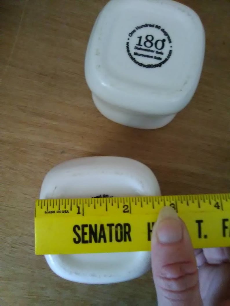 May include: Two white ceramic square bowls with rounded edges. The bottom of one bowl has a black sticker with the text "One Hundred 80 degrees Dishwasher Safe Microwave Safe". The other bowl is being measured with a yellow ruler that reads "SENATOR H T. F" and has markings in inches.