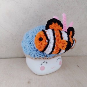 May include: A blue crocheted mug cosy with a crocheted orange and white clownfish on top. The mug cosy is on a white mug with a pink smiling face.
