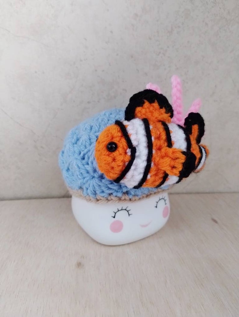 May include: A blue crocheted mug cosy with a crocheted orange and white clownfish on top. The mug cosy is on a white mug with a pink smiling face.