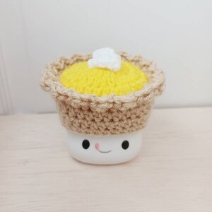 May include: A white marshmallow with a crocheted pie crust hat. The pie crust is brown and the filling is yellow with a white dollop of whipped cream on top. The marshmallow has a smiling face.