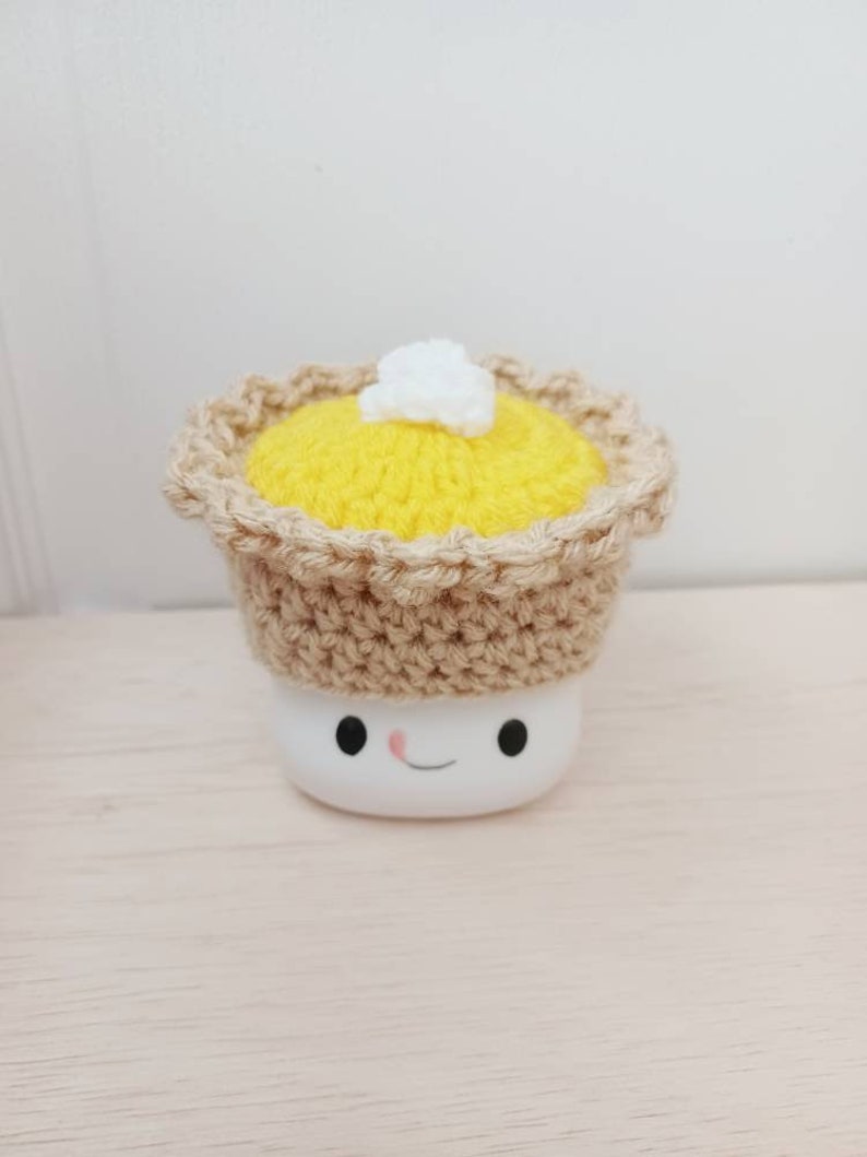 May include: A white marshmallow with a crocheted pie crust hat. The pie crust is brown and the filling is yellow with a white dollop of whipped cream on top. The marshmallow has a smiling face.