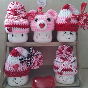 May include: Six white ceramic mugs with different crocheted hats in pink, red, and white with hearts and bows. The mugs have painted faces and are arranged on a wooden shelf. A red heart is in the foreground.