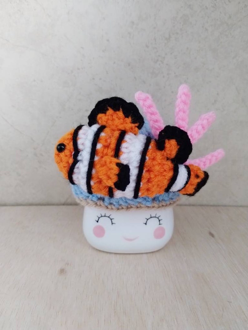May include: A crocheted clownfish with orange, white, and black stripes sits on top of a white mug with a smiling face. The clownfish has a pink coral attached to it.