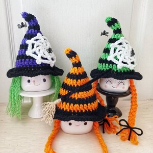 May include: Three ceramic mugs with crocheted witch hats and yarn hair. The hats are black, green, and orange with white spiderwebs. The mugs have painted faces and are on white pedestals.