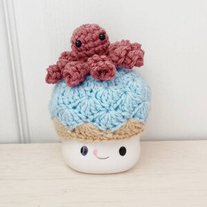 May include: A white ceramic mug with a smiling face, wearing a blue and tan crocheted hat with a pink crocheted octopus on top.