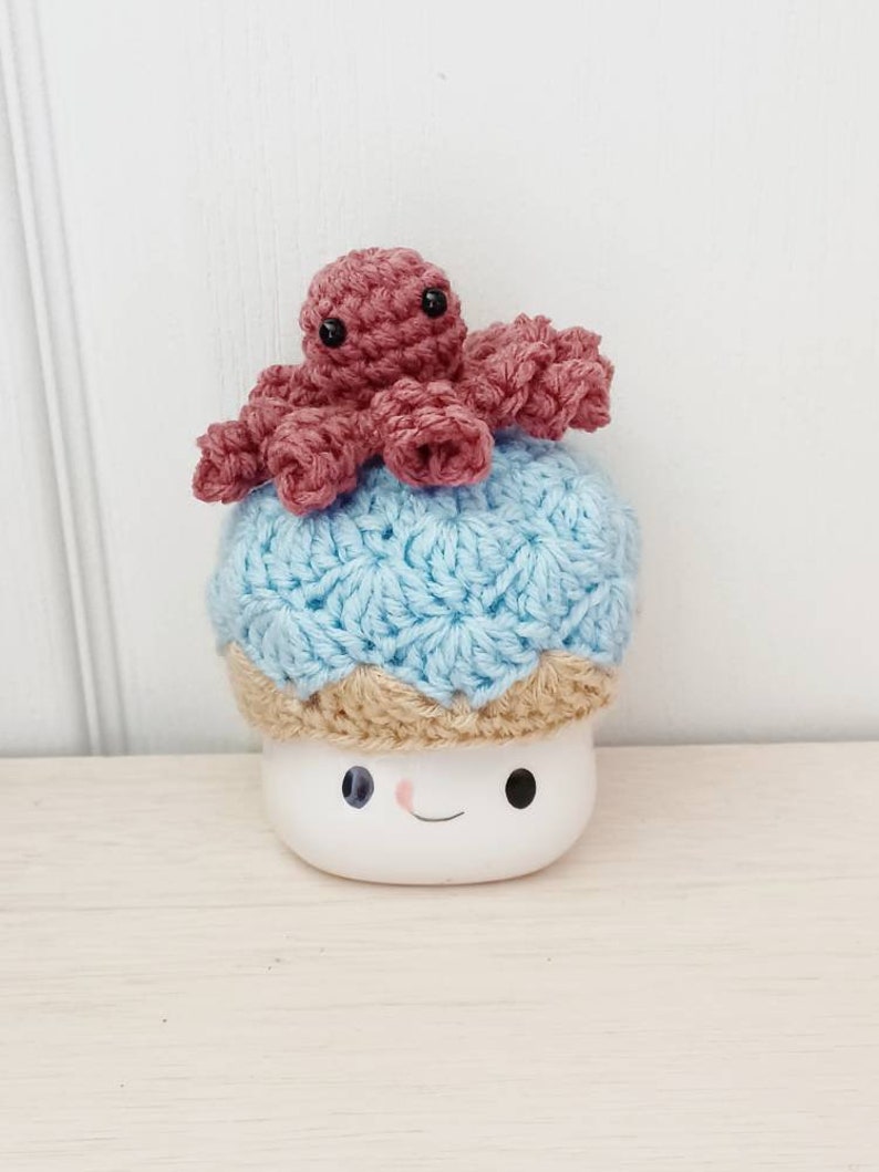 May include: A white ceramic mug with a smiling face, wearing a blue and tan crocheted hat with a pink crocheted octopus on top.