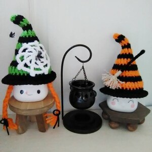 May include: Two crocheted witch hats with marshmallow faces and orange yarn hair. The hats are decorated with black and white stripes, a spider web, and a broom. The hats are sitting on small wooden stools. A black cauldron hangs from a metal stand.