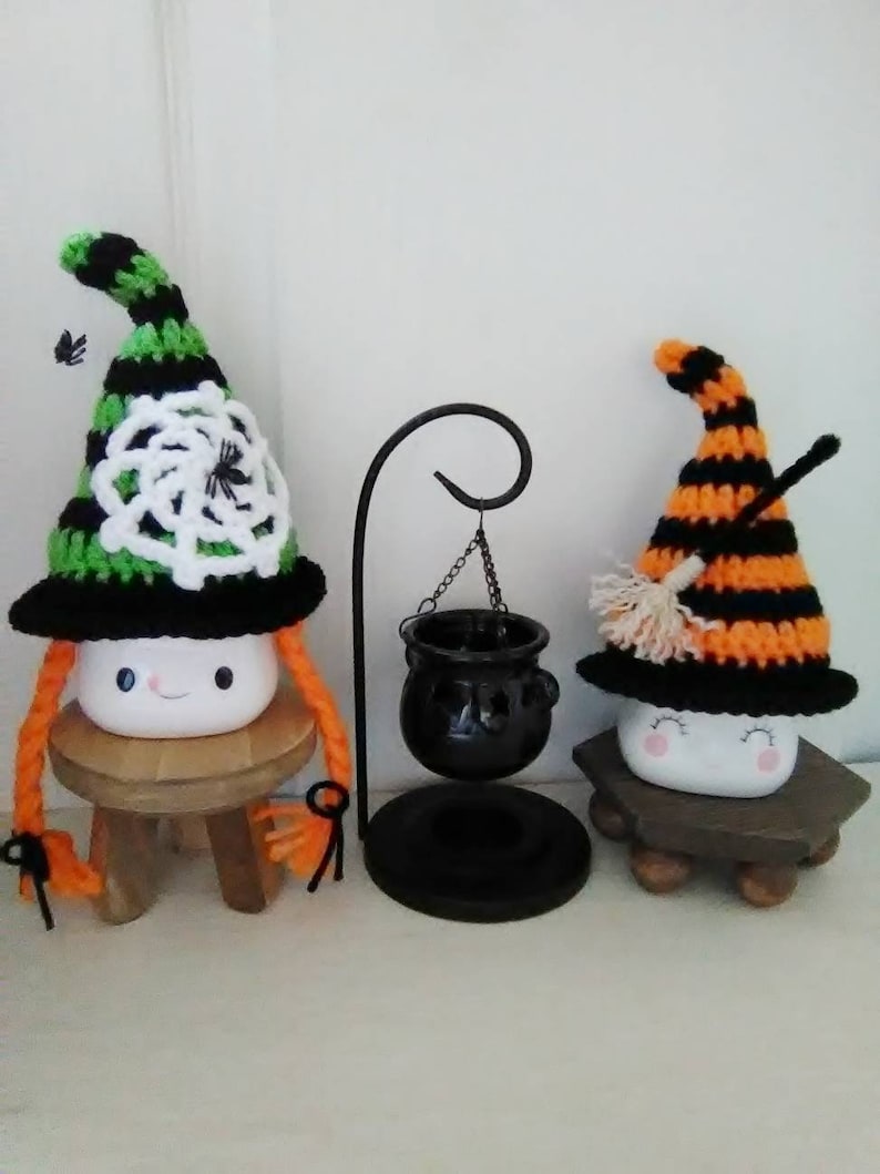 May include: Two crocheted witch hats with marshmallow faces and orange yarn hair. The hats are decorated with black and white stripes, a spider web, and a broom. The hats are sitting on small wooden stools. A black cauldron hangs from a metal stand.