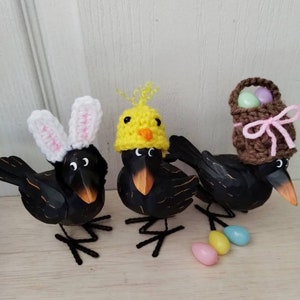 K&ouml;nnte beinhalten: Drei schwarze Vogelfiguren mit Osterh&uuml;ten. Ein Vogel tr&auml;gt Hasenohren, ein anderer einen K&uuml;kenhut und der dritte einen Korbhut. Pastellfarbene Eier sind vor den V&ouml;geln verstreut. Die Figuren stehen auf einer hellen Holzoberfl&auml;che.