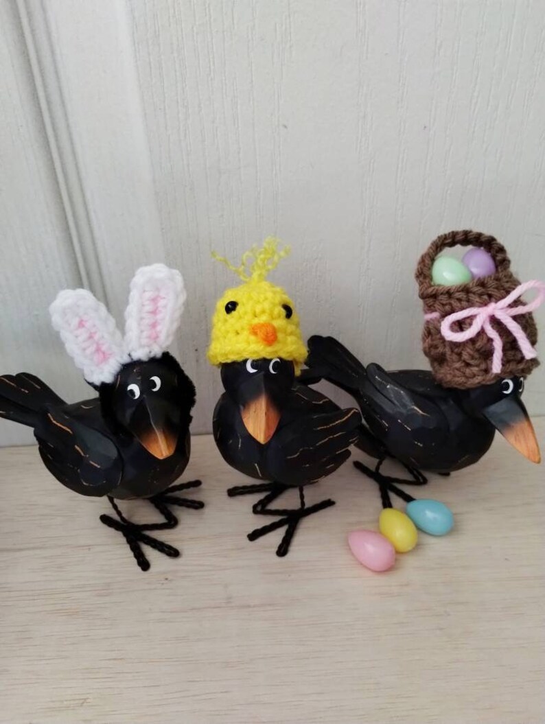 K&ouml;nnte beinhalten: Drei schwarze Vogelfiguren mit Osterh&uuml;ten. Ein Vogel tr&auml;gt Hasenohren, ein anderer einen K&uuml;kenhut und der dritte einen Korbhut. Pastellfarbene Eier sind vor den V&ouml;geln verstreut. Die Figuren stehen auf einer hellen Holzoberfl&auml;che.