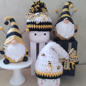 May include: Three gnomes wearing crocheted bee-themed hats. The hats are white with black and yellow stripes and have bee embellishments. The gnomes are made of ceramic and have white beards.