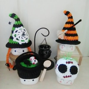 May include: Three Halloween-themed marshmallow mugs with crocheted hats. The first mug has a green and black witch hat with a white spiderweb design. The second mug has an orange and black witch hat with a black broom. The third mug has a white skull hat with black eyes and a stitched mouth.