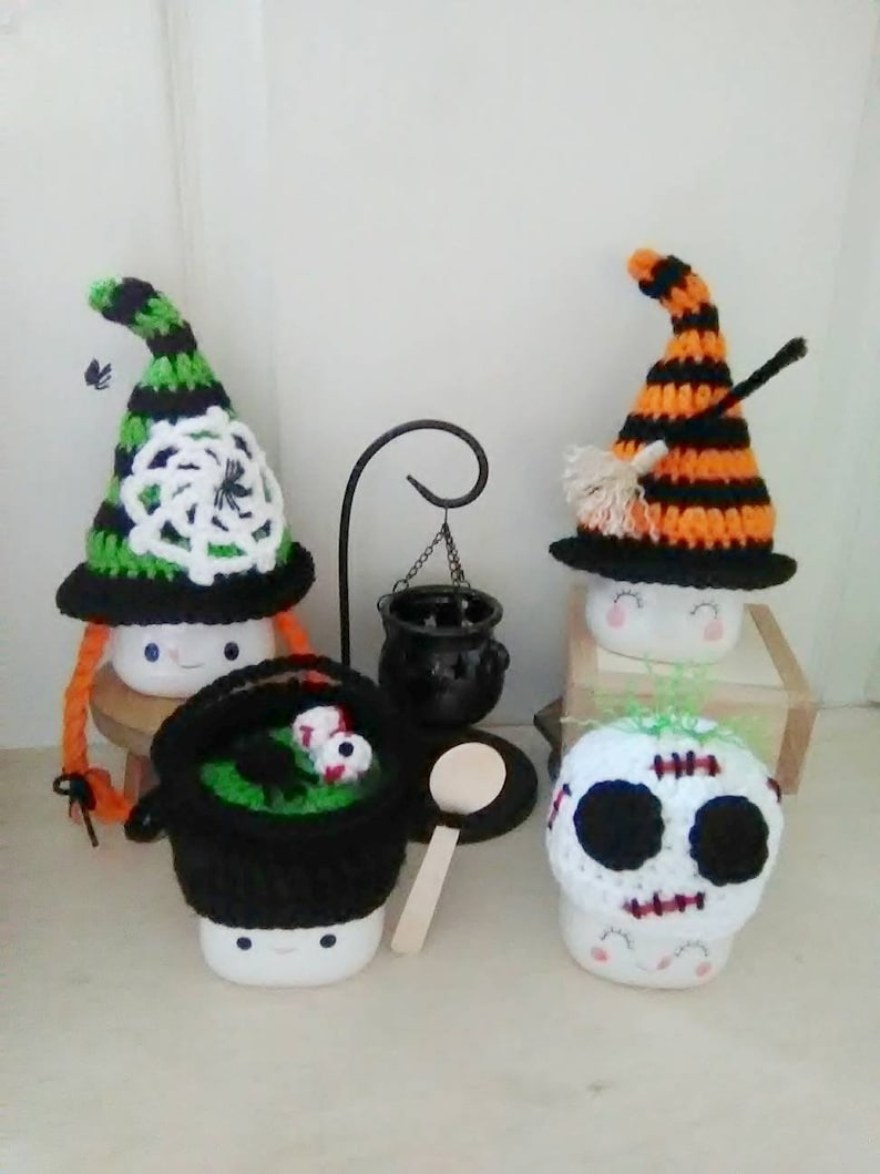 May include: Three Halloween-themed marshmallow mugs with crocheted hats. The first mug has a green and black witch hat with a white spiderweb design. The second mug has an orange and black witch hat with a black broom. The third mug has a white skull hat with black eyes and a stitched mouth.