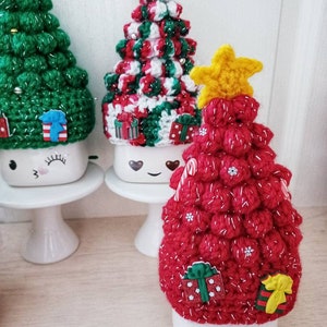 May include: Three ceramic mugs with crocheted Christmas tree toppers. The mugs have a white background with a black smiley face and eyelashes. The tree toppers are red, green, and white with a yellow star on top.