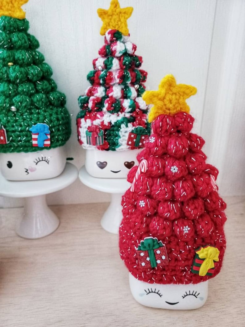 May include: Three ceramic mugs with crocheted Christmas tree toppers. The mugs have a white background with a black smiley face and eyelashes. The tree toppers are red, green, and white with a yellow star on top.
