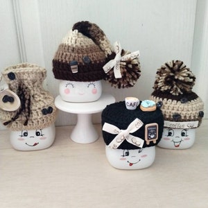 May include: Four white ceramic mugs with different crocheted hats. The mugs have painted faces and are decorated with coffee-themed details. The hats are brown, black, and beige with pom-poms.