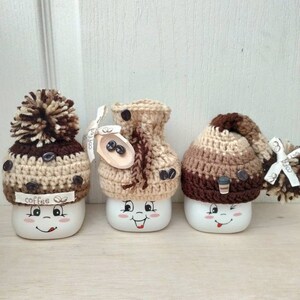May include: Three white ceramic mugs with happy faces, each wearing a different brown and beige crocheted hat. The hats have coffee bean details and one has a pom-pom.