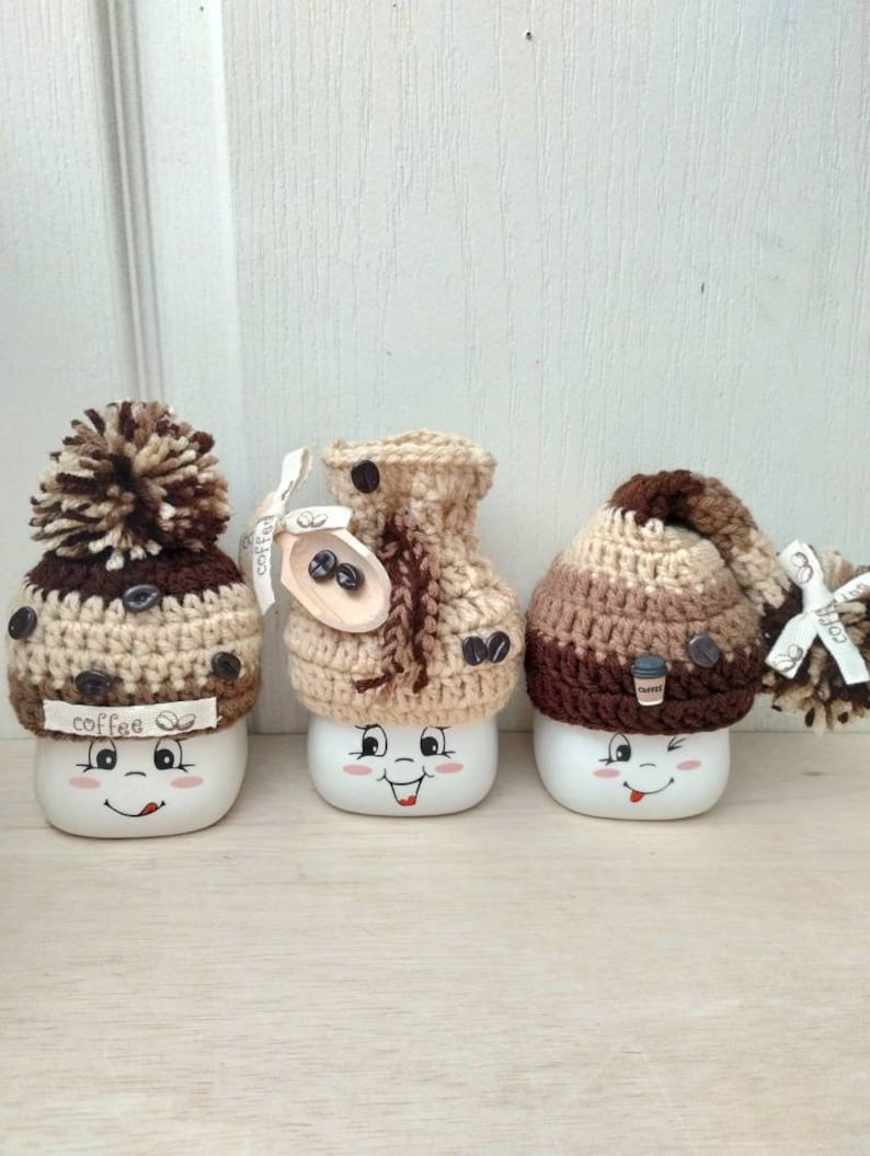 May include: Three white ceramic mugs with happy faces, each wearing a different brown and beige crocheted hat. The hats have coffee bean details and one has a pom-pom.