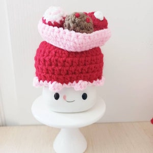 May include: A crocheted amigurumi cake with pink and red frosting and a brown chocolate topping. The cake is sitting on a white cake stand.