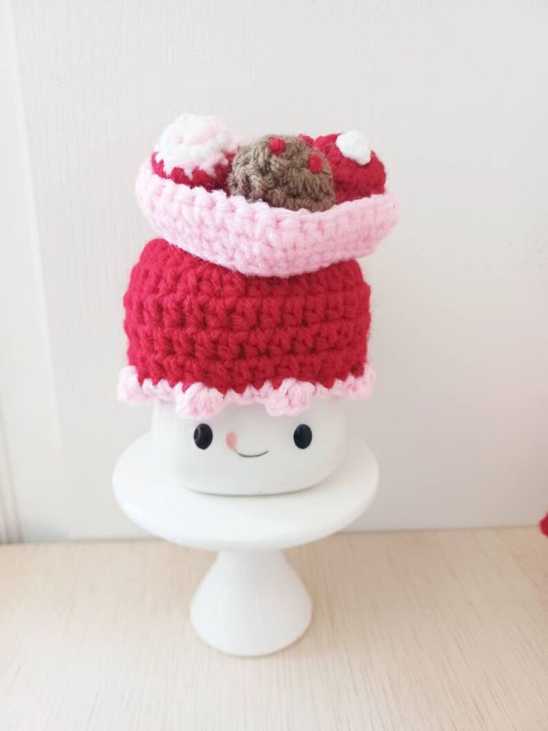 May include: A crocheted amigurumi cake with pink and red frosting and a brown chocolate topping. The cake is sitting on a white cake stand.