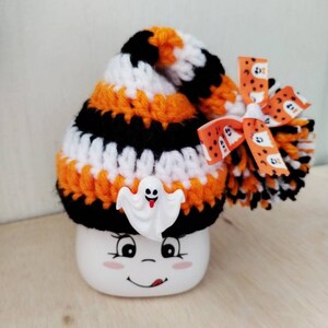 May include: A white ceramic mug with a smiling face and a black, orange, and white striped crocheted hat with a ghost charm and a bow with a ghost pattern.