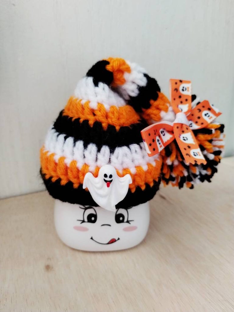 May include: A white ceramic mug with a smiling face and a black, orange, and white striped crocheted hat with a ghost charm and a bow with a ghost pattern.