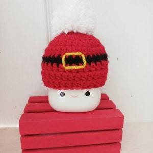Op de afbeelding: Een witte keramische mok met een rood-zwarte gehaakte kerstmanmuts. De mok heeft een glimlachend gezicht.