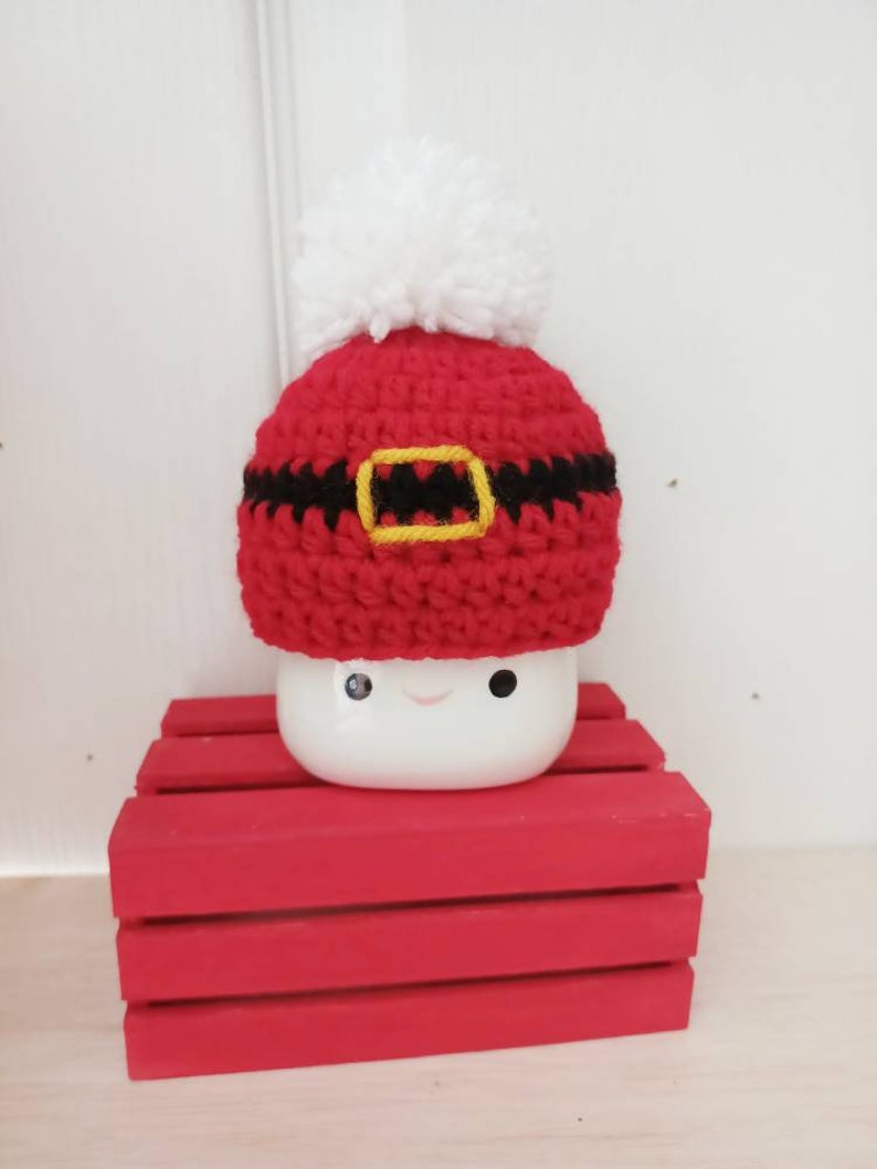 Op de afbeelding: Een witte keramische mok met een rood-zwarte gehaakte kerstmanmuts. De mok heeft een glimlachend gezicht.