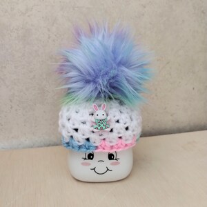 May include: A white crocheted hat with a blue, pink, and green stripe pattern. The hat has a fluffy blue pom-pom on top and a small white bunny charm with pink polka dots.