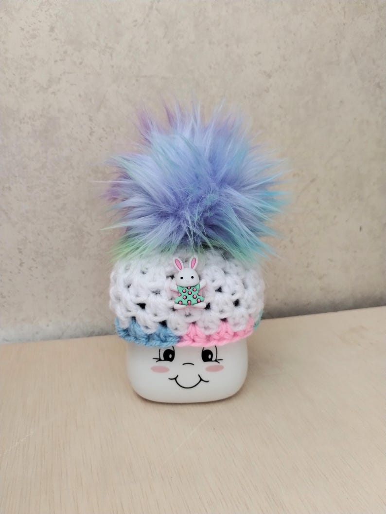 May include: A white crocheted hat with a blue, pink, and green stripe pattern. The hat has a fluffy blue pom-pom on top and a small white bunny charm with pink polka dots.