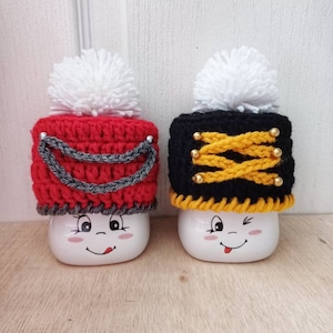 May include: Two white ceramic mugs with cute faces wearing crocheted hats. The hats are red and black with white pom poms on top. The red hat has a silver trim and the black hat has gold buttons.