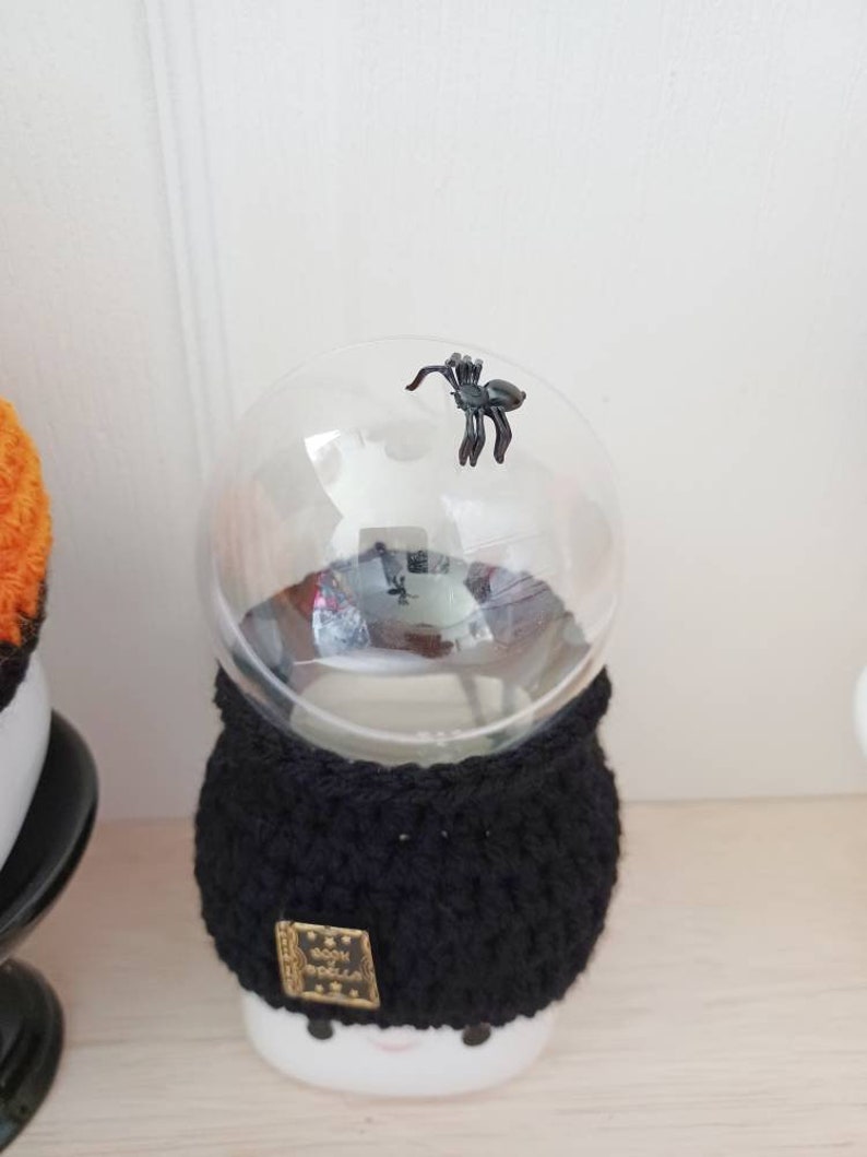 K&ouml;nnte beinhalten: Ein Halloween-Dekorationsobjekt mit einer durchsichtigen Glaskugel, auf der sich eine kleine schwarze Spinne befindet. Die Kugel sitzt auf einem wei&szlig;en Sockel mit einer schwarzen geh&auml;kelten H&uuml;lle. Ein kleines goldfarbenes Etikett ist an der H&uuml;lle befestigt.
