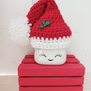 Op de afbeelding: Een witte keramische mok met een glimlachend gezicht en wimpers, die een rode gehaakte kerstmanmuts draagt met een witte pompon en een groene hulstversiering. De mok staat op een rode houten kist.