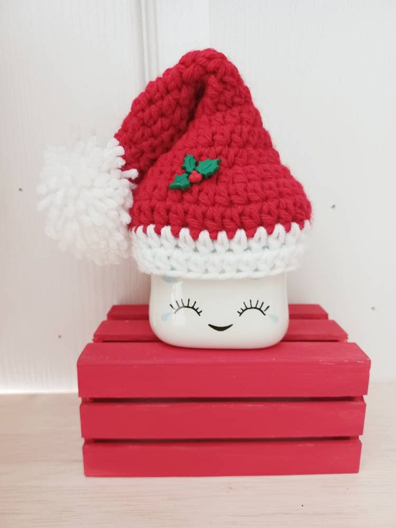 Op de afbeelding: Een witte keramische mok met een glimlachend gezicht en wimpers, die een rode gehaakte kerstmanmuts draagt met een witte pompon en een groene hulstversiering. De mok staat op een rode houten kist.