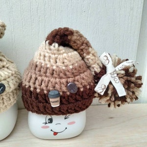 May include: A white ceramic mug with a brown, beige, and dark brown crocheted hat with a pom-pom and a small coffee cup charm. The mug has a cartoon face with a winking eye and a tongue sticking out.