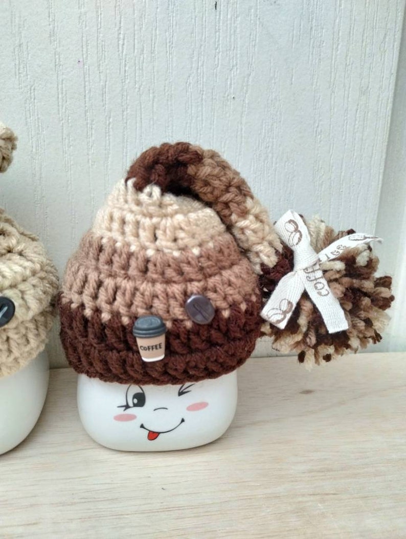 May include: A white ceramic mug with a brown, beige, and dark brown crocheted hat with a pom-pom and a small coffee cup charm. The mug has a cartoon face with a winking eye and a tongue sticking out.