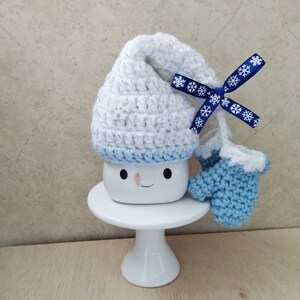 K&ouml;nnte beinhalten: Eine wei&szlig;e Keramiktasse mit einem l&auml;chelnden Gesicht, die einen wei&szlig;en H&auml;kelhut mit einem blauen Band und blauen H&auml;kelhandschuhen tr&auml;gt. Die Tasse steht auf einem wei&szlig;en Kuchenst&auml;nder.
