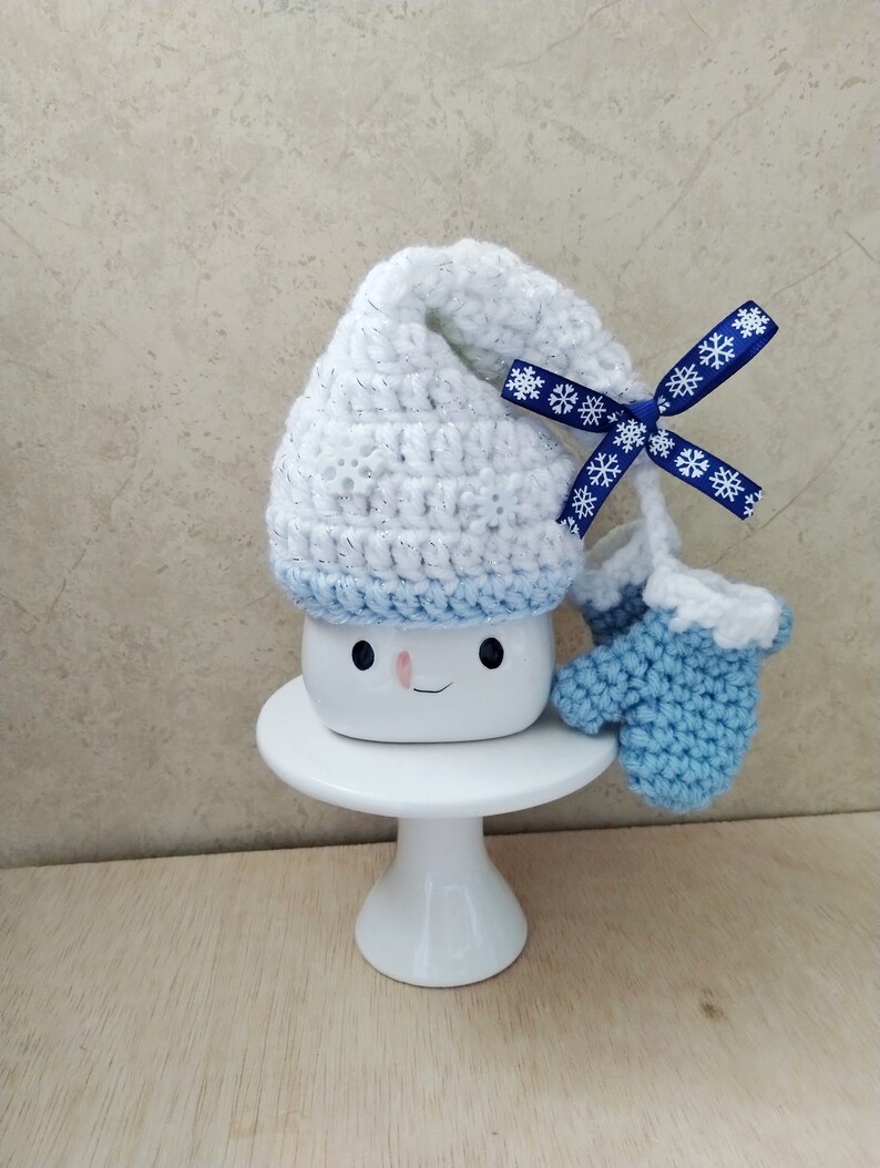 K&ouml;nnte beinhalten: Eine wei&szlig;e Keramiktasse mit einem l&auml;chelnden Gesicht, die einen wei&szlig;en H&auml;kelhut mit einem blauen Band und blauen H&auml;kelhandschuhen tr&auml;gt. Die Tasse steht auf einem wei&szlig;en Kuchenst&auml;nder.