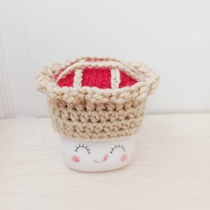 May include: A white ceramic mug with a smiling face and closed eyes, wearing a crocheted pie hat with a red filling and a tan crust.