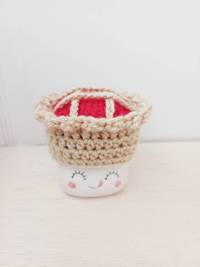 May include: A white ceramic mug with a smiling face and closed eyes, wearing a crocheted pie hat with a red filling and a tan crust.