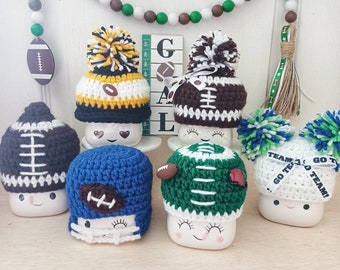 May include: Six white ceramic mugs with crocheted hats in various colors and designs, including green, blue, brown, and black. The hats feature football stitching and some have pom-poms. The mugs have happy faces and some have the text "Go Team!" on them.