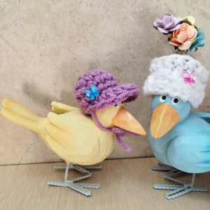 Op de afbeelding: Twee houten vogelbeeldjes, een gele met een paarse gebreide hoed en de andere blauwe met een witte gebreide hoed en een bloemversiering. Beide vogels hebben wit geschilderde voeten.