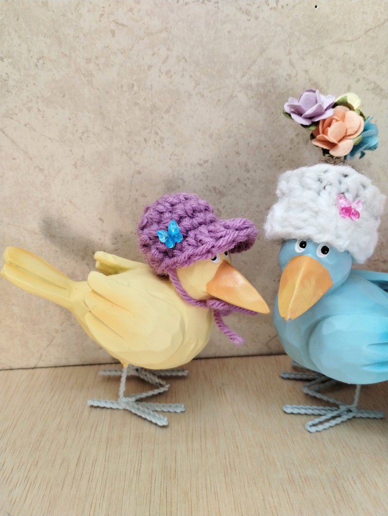 Op de afbeelding: Twee houten vogelbeeldjes, een gele met een paarse gebreide hoed en de andere blauwe met een witte gebreide hoed en een bloemversiering. Beide vogels hebben wit geschilderde voeten.