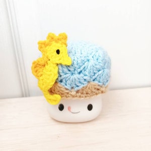 May include: A white ceramic marshmallow with a blue and brown crocheted hat and a yellow crocheted seahorse on top.